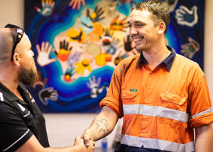 2 men standing smiling and shaking hands in front of aboriginal art