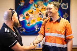 2 men shaking hands and smiling standing in front of aboriginal art